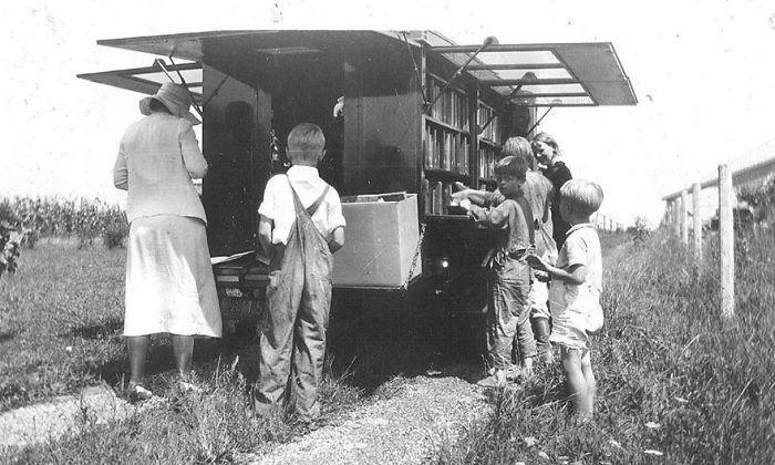Children and an adult browsing books at a vintage bookmobile, an early form of libraries-on-wheels in a rural setting.