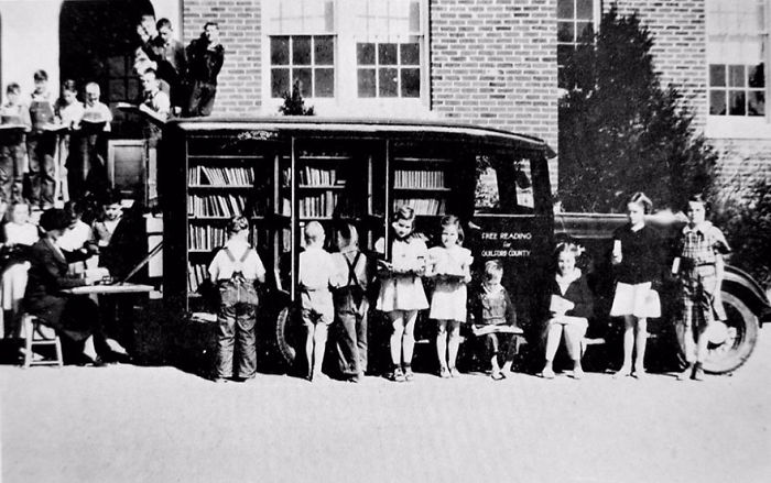 Children reading books outside a vintage bookmobile library-on-wheels parked near a brick building in a black and white photo.