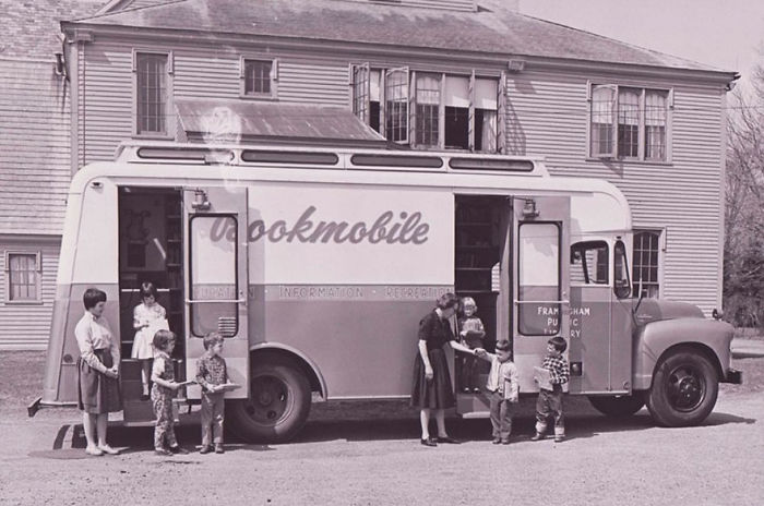 Vintage bookmobile parked outside a building, with children and adults boarding and receiving books from the mobile library.