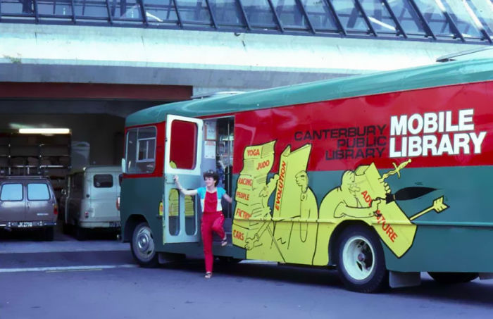 A woman stepping out of a colorful Canterbury Public Library mobile library bookmobile parked outside a building.