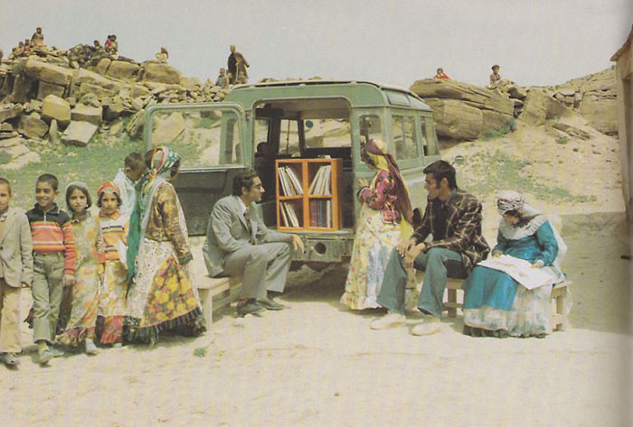 Children and adults gathered around a bookmobile in a rural area with shelves of books visible inside the vehicle.