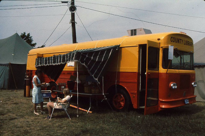 Vintage county library bookmobile with yellow and orange exterior, serving books outdoors under a striped awning on grass.