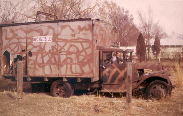 Old bookmobile vehicle with camouflage paint parked in a rural area, showcasing libraries on wheels from the past.
