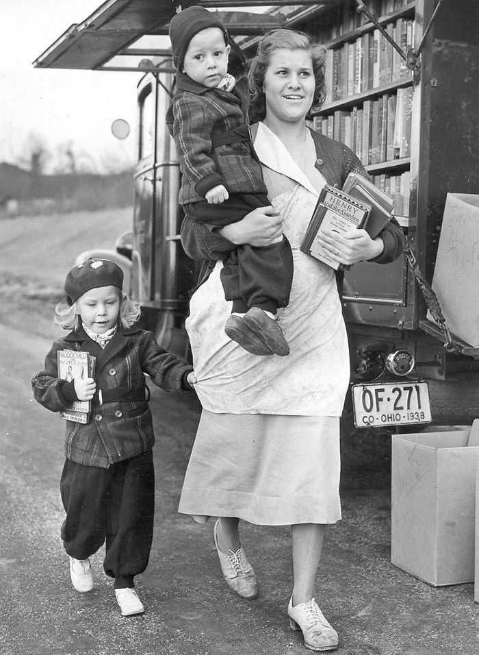 Woman carrying a child and holding books next to a vintage bookmobile, with a young girl holding a book nearby.