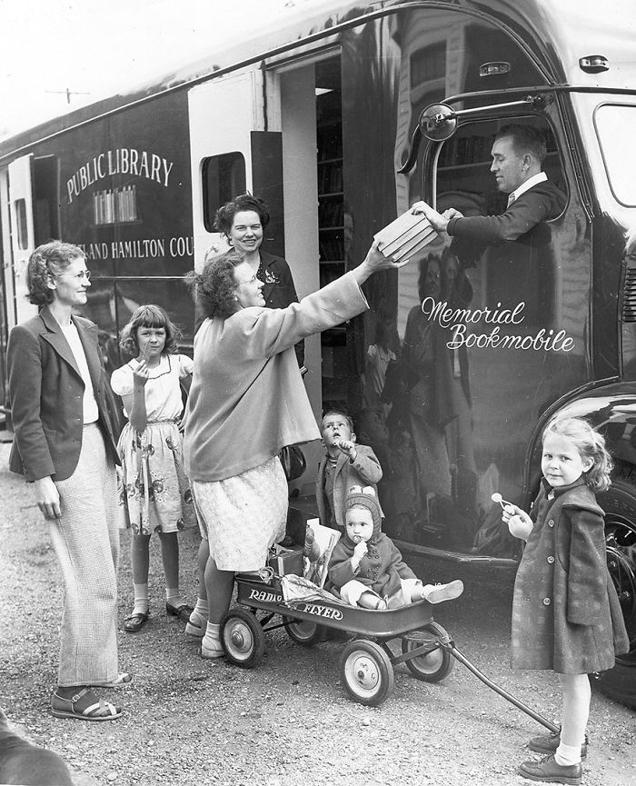 Woman receiving books from a public library bookmobile while children and adults wait nearby in a vintage black and white photo.