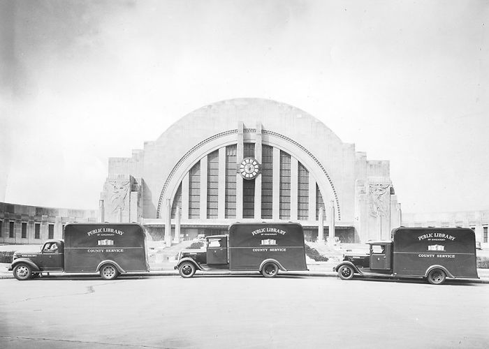 Three vintage bookmobiles parked in front of a large public library building, showcasing libraries-on-wheels history.