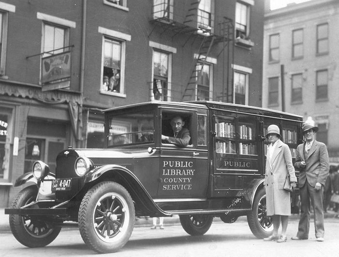Vintage public library bookmobile parked on a city street with man driving and two people standing nearby.