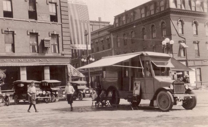 Vintage black and white photo of a bookmobile parked in a town square with people nearby in early 20th century setting.