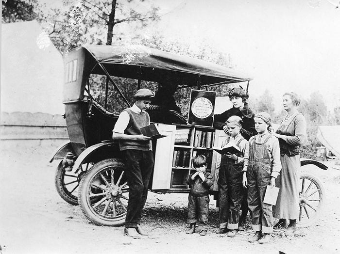 Black and white photo of a bookmobile with people reading books outside the mobile library in a vintage setting.