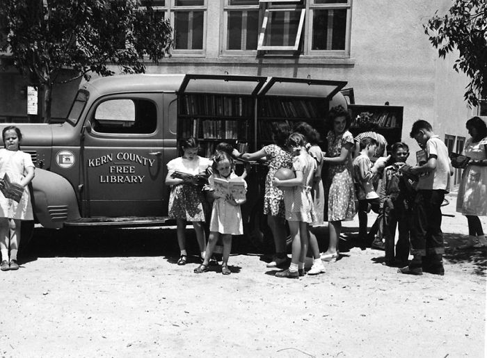 Vintage black and white photo of children gathered around a Kern County free library bookmobile reading books outdoors.