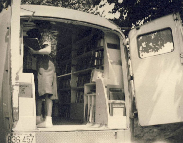 Woman inside a vintage bookmobile browsing books on shelves in a mobile library with open rear doors.