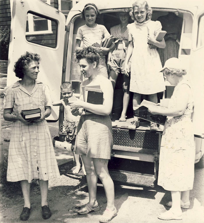 Group of women and children with books by an open bookmobile, showcasing libraries on wheels in a vintage black and white photo.