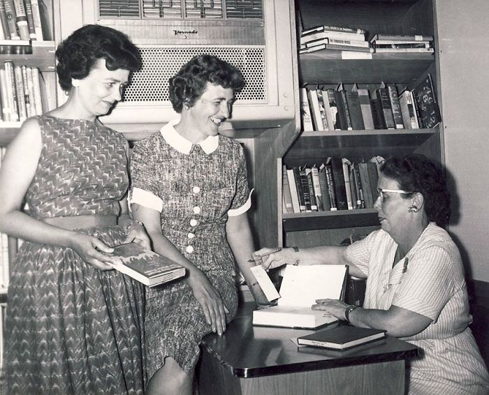 Three women interacting inside a vintage bookmobile, showcasing rare libraries on wheels with shelves of books in a black and white photo.