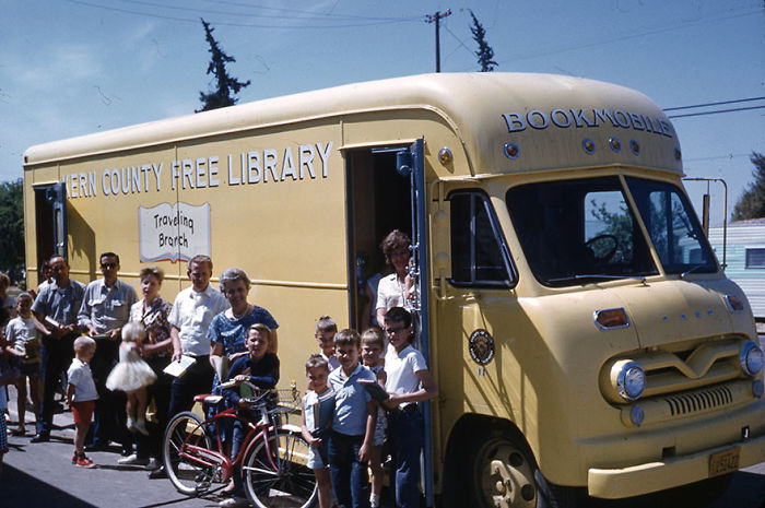A group of children and adults gathered around a vintage yellow bookmobile from a free library traveling branch.