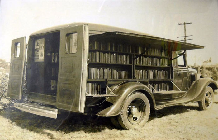 Vintage bookmobile with open side panels displaying shelves filled with books, showcasing libraries-on-wheels in early 20th century.