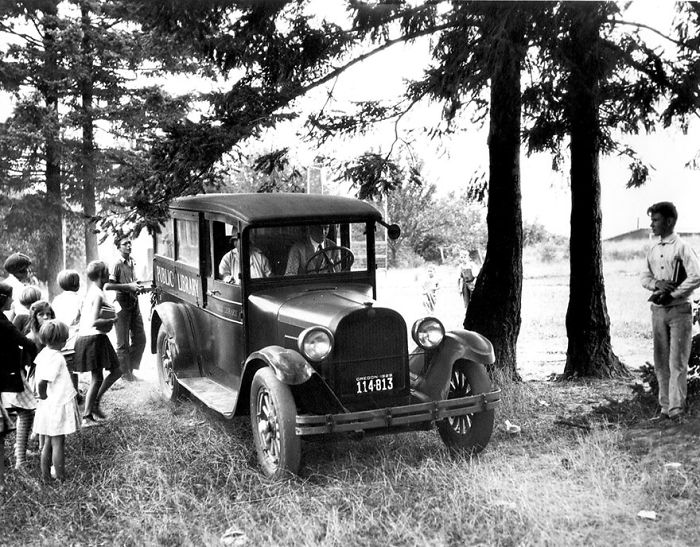 Vintage bookmobile parked under trees, with children gathered around, illustrating libraries on wheels before Amazon.