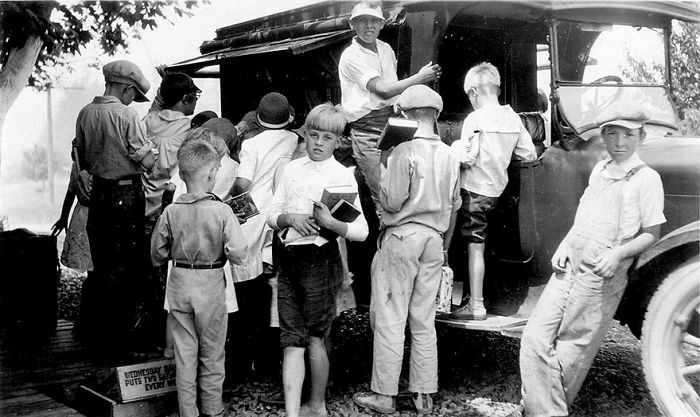 Children gathered around a vintage bookmobile, exploring books from a mobile library vehicle in a historical setting.