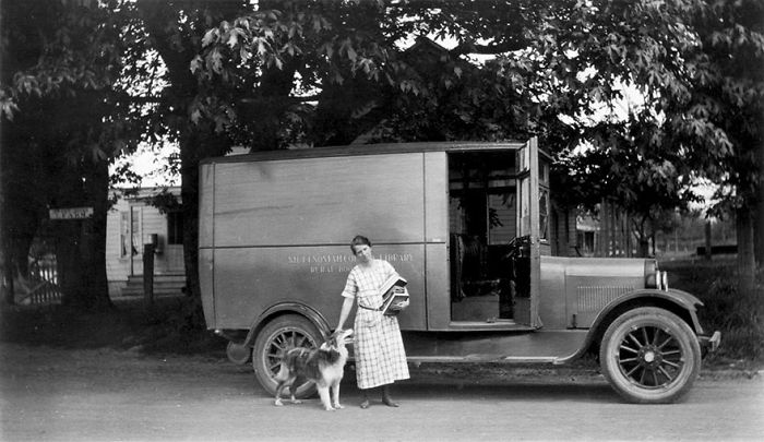 Woman with dog standing beside a vintage bookmobile library vehicle under large trees on a rural street.