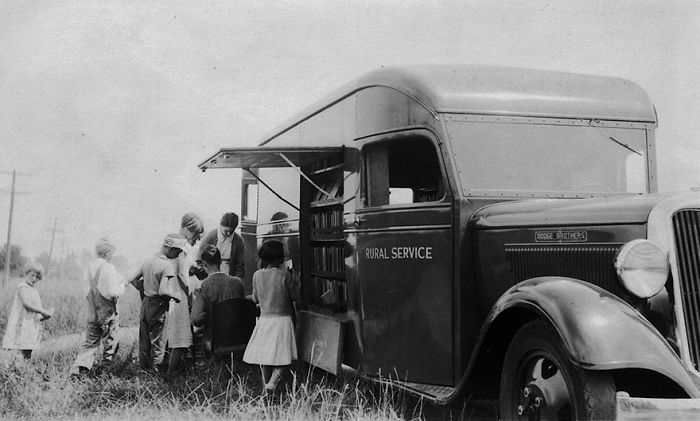 Vintage bookmobile parked in a rural area with children gathered around, showcasing libraries-on-wheels before Amazon's era.