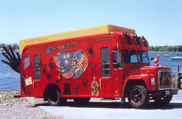 Red bookmobile decorated with colorful art, parked by the waterfront, representing a mobile library on wheels.