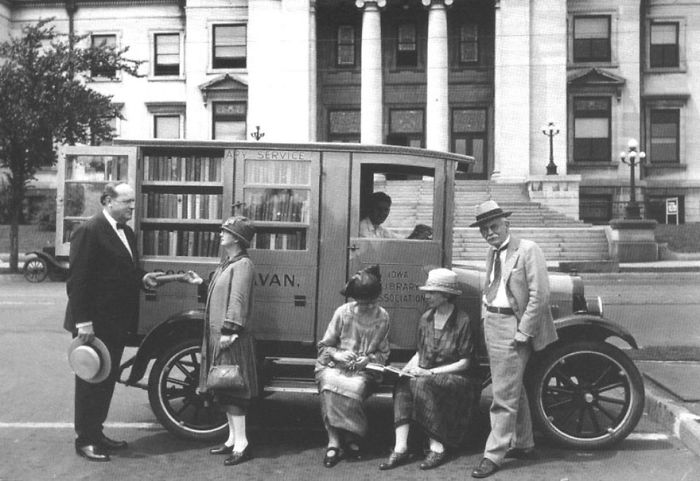 Vintage bookmobile parked outside a building with people reading and interacting around the libraries-on-wheels vehicle.