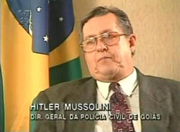 Man in a suit and tie named with a funny name on TV, Brazilian flag in the background.