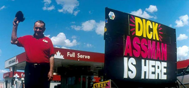Man in front of a gas station sign with "D**k Assman is here," showcasing funny names.