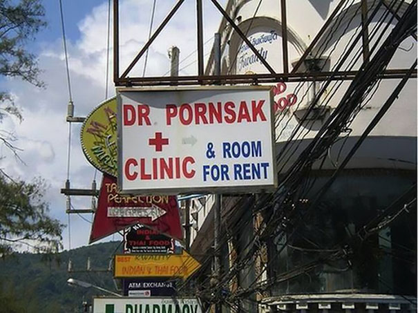 Funny name sign: "Dr. Pornsak Clinic & Room for Rent" on a busy street with other colorful signs.