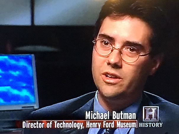 Man in glasses and suit, identified as Michael Butman, with a funny name, speaking on History channel while at a museum.
