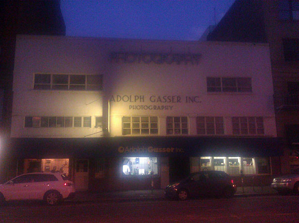 Storefront at night with a sign displaying a funny name, featuring parked cars on the street.
