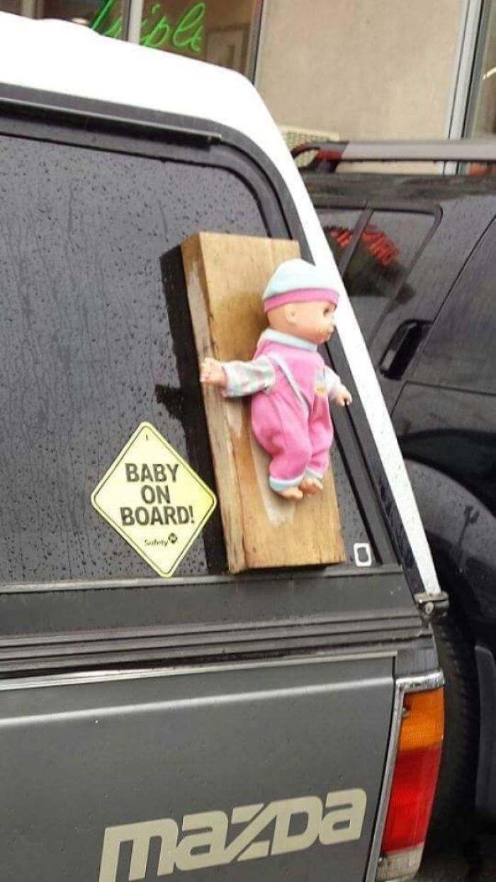A doll humorously strapped to a truck with a "Baby on Board" sign, showcasing dads' jokes taken to another level.
