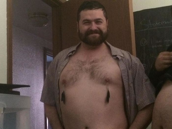 Dad jokes gone wild: Man humorously sports feather earrings on his chest while smiling indoors.