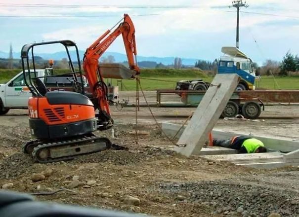 Excavator lifting concrete slab while worker in neon vest lies in ditch, illustrating reasons women may live longer than men.