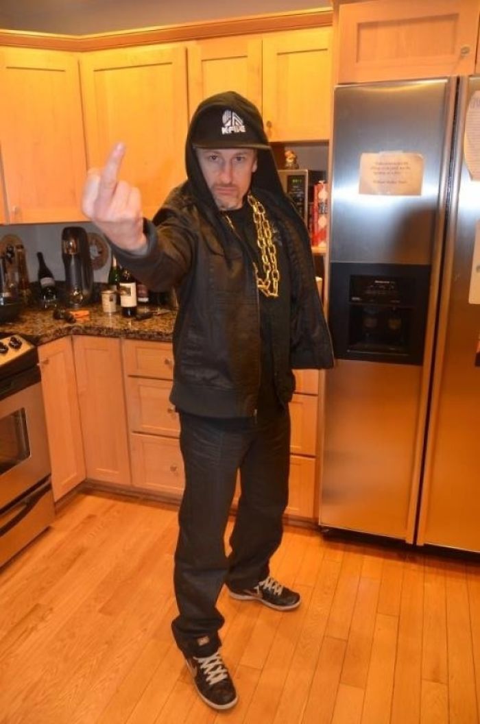 Dad in a kitchen wearing a hoodie, hat, and gold chain, humorously posing with finger up.
