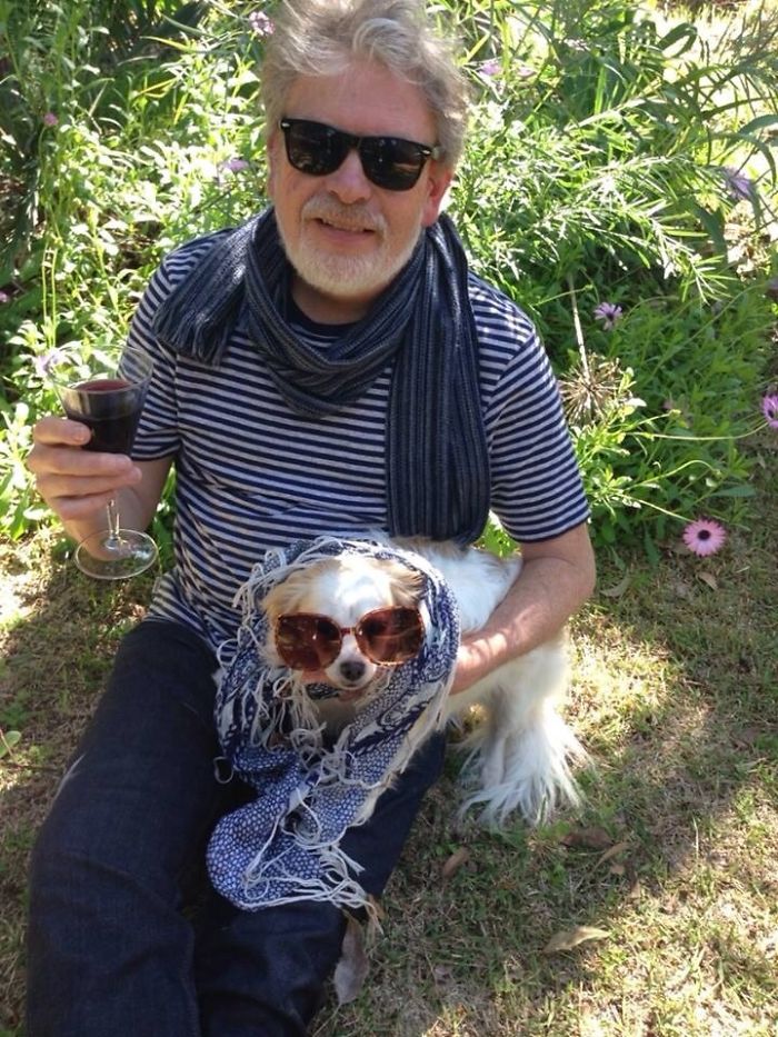 Dad with sunglasses and scarf, holding a drink, sharing a funny moment with a dog also in sunglasses.