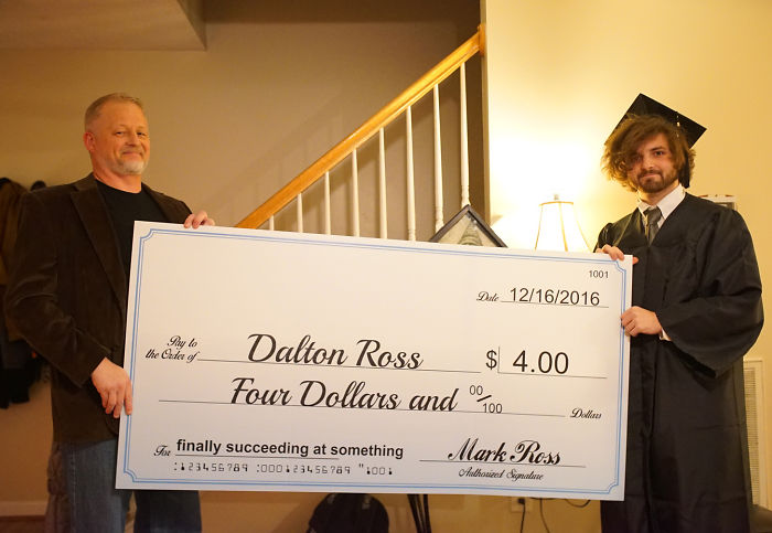 Dad holding oversized check with dollar amount as a joke; son in graduation attire smiling.