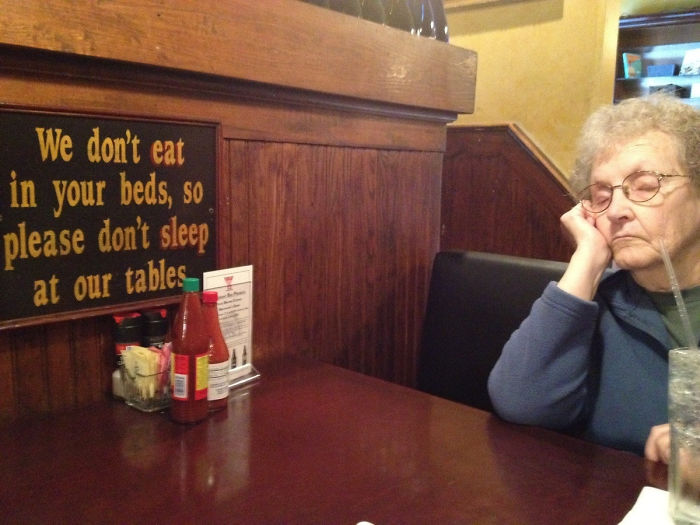 Elderly woman asleep at a restaurant table next to a humorous sign, embodying people who don't care.