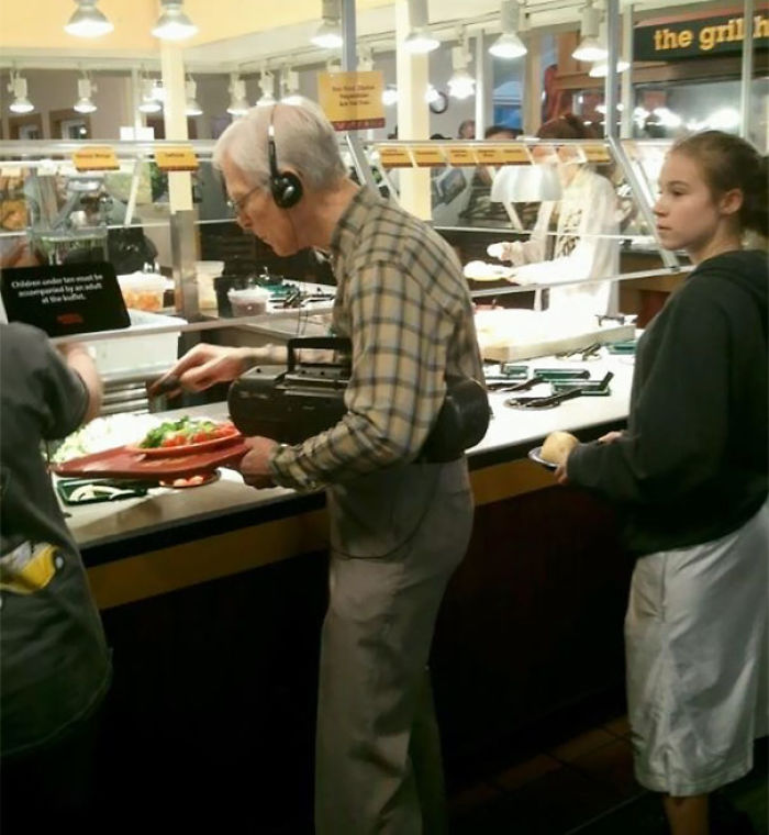 Elderly man listening to a boombox while serving food at a buffet, showing indifference.