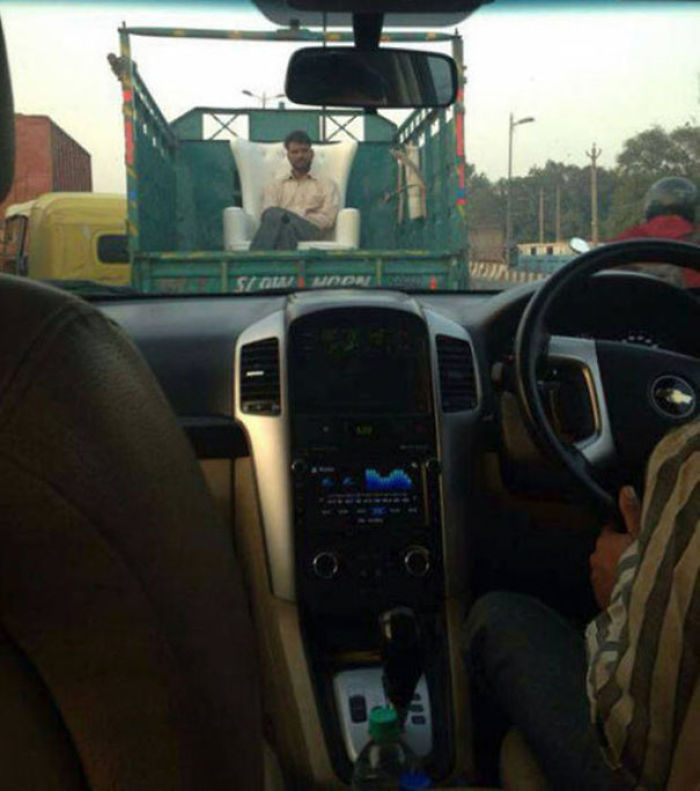 Man sitting in an armchair on a moving truck, viewed from a car interior, depicting indifference to safety.