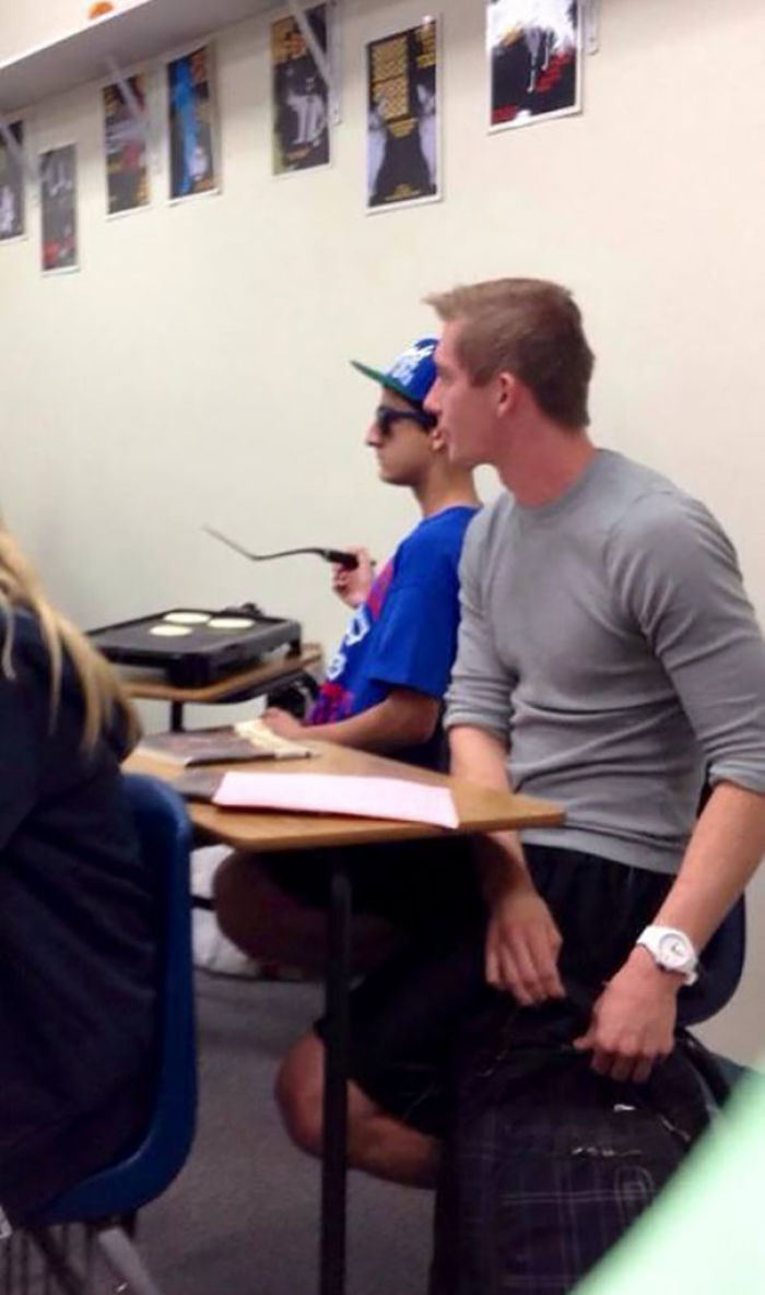 Two students in a classroom setting, with one wearing sunglasses and a cap, appear unbothered while cooking pancakes.