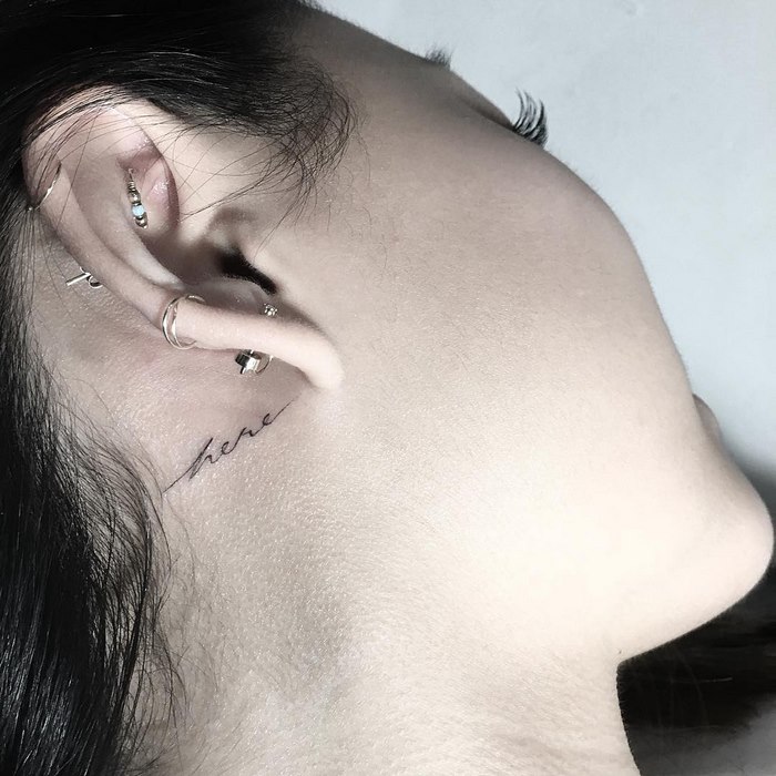 Delicate typographic tattoo "here" behind ear, showcasing fine line artistry by Léo Gavaggio.