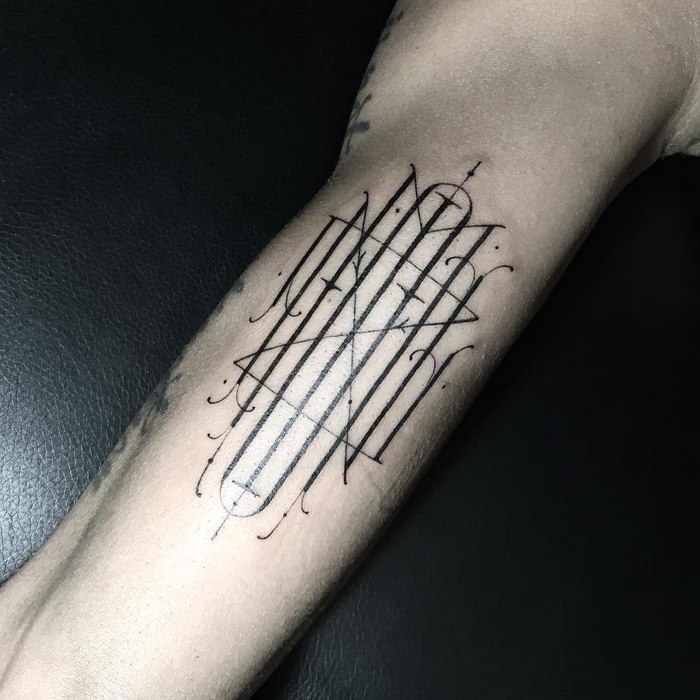 Delicate black typographic tattoo on a forearm, featuring intricate lines and patterns.