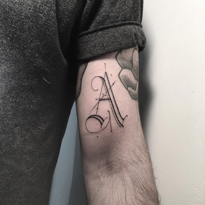 Delicate typographic tattoo of an ornate letter on a person's upper arm.