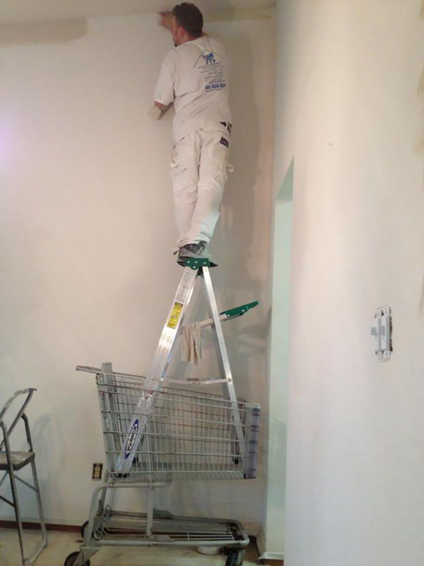 Man balancing on a ladder inside a shopping cart while painting a wall, demonstrating risky behavior.