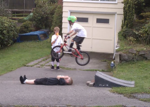 Boy on bike jumps over another lying on the ground, with a friend watching.