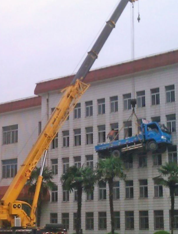 Crane lifting a blue truck near a building; a man stands on the truck, illustrating risky actions related to lifespan differences.