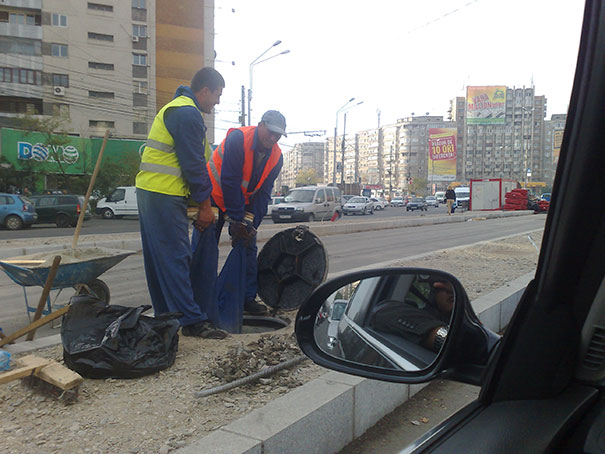 Two men in safety vests working on a roadside construction site, lifting a heavy object near a busy street.