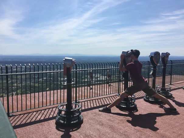 Tall person stretching legs awkwardly to use a coin-operated binocular viewer at a scenic overlook on a sunny day.
