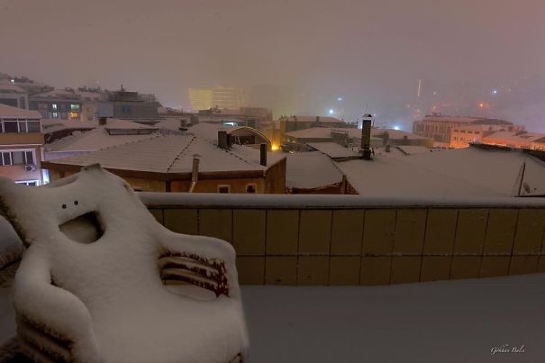 Snow-covered chair on balcony showing funny pareidolia face in a nighttime cityscape with rooftops in the background