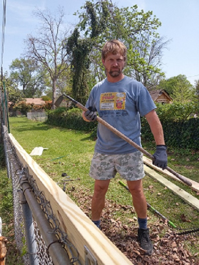 Dad in a humorous t-shirt, holding a shovel, standing by a wooden fence in a backyard.